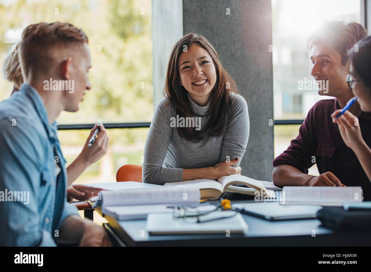Sorridente giovane donna seduta a tavola con i compagni di classe nella libreria. Un gruppo di giovani felici gli studenti a studiare in biblioteca. Foto Stock Sorridente giovane donna seduta a tavola con i compagni di classe nella libreria. Un gruppo di giovani felici gli studenti a studiare in biblioteca. Foto Stock