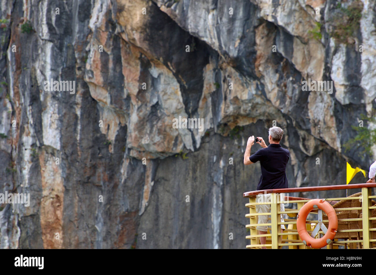 Turista occidentale tiene iPhone Foto di Halong Bay, Vietnam mentre si sta in piedi sul ponte superiore della barca con una parete rocciosa isola di sfondo Foto Stock