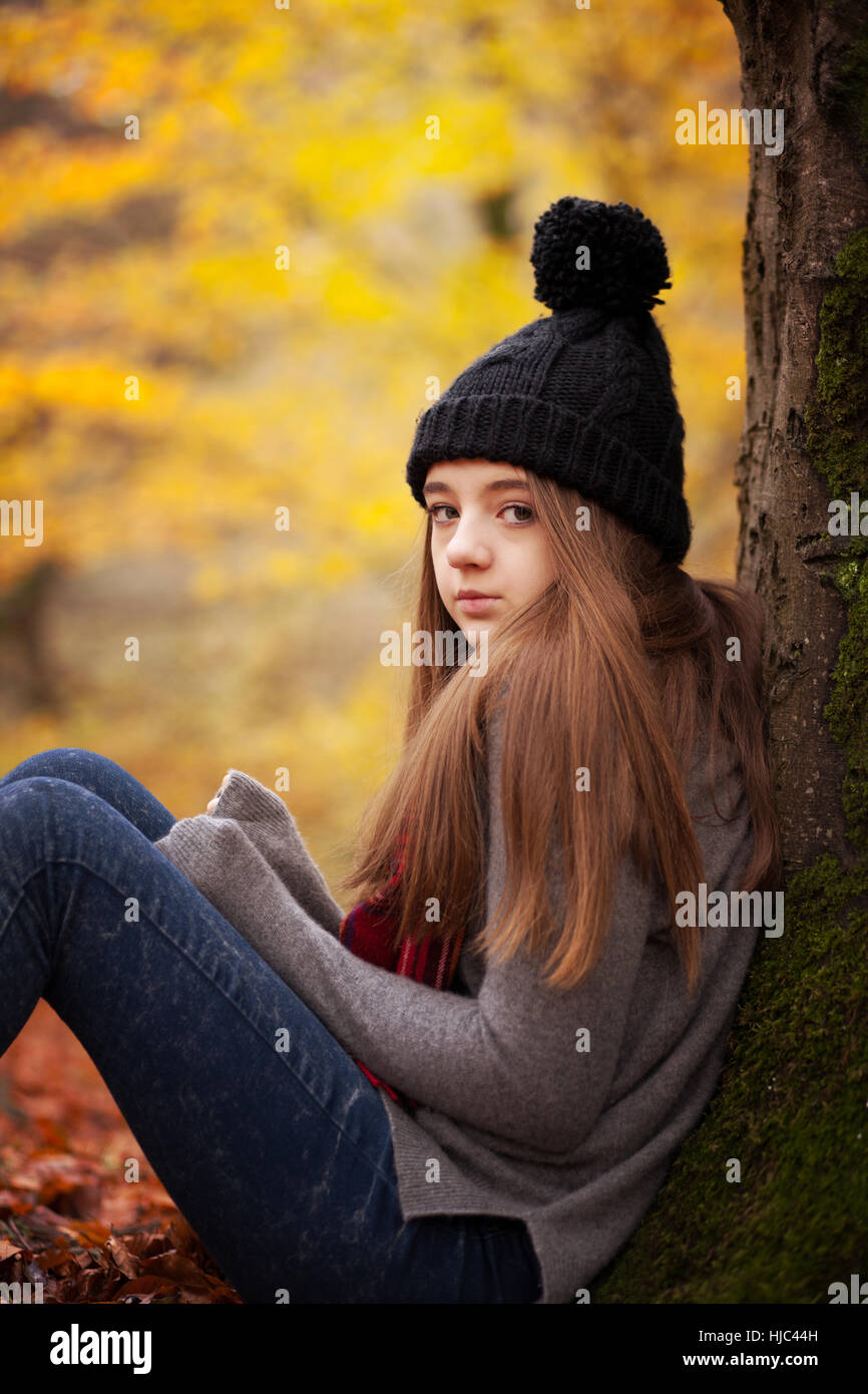 Ragazza adolescente che indossa un cappello di lana seduto accanto a un albero con colori autunnali in background Foto Stock