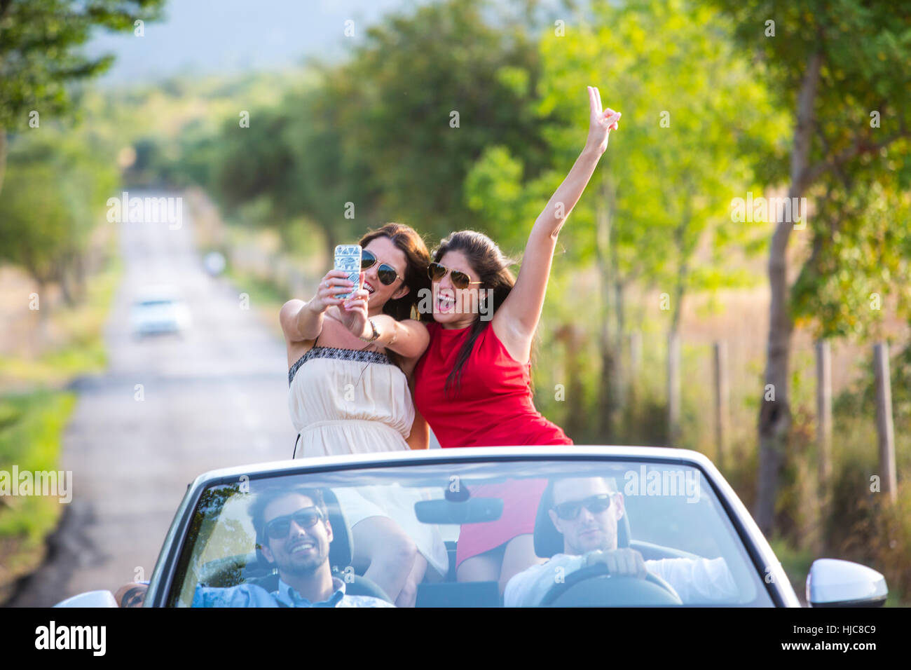 Due giovani amici di sesso femminile prendendo selfie durante la guida su strada rurale Nel convertibile, Maiorca, SPAGNA Foto Stock