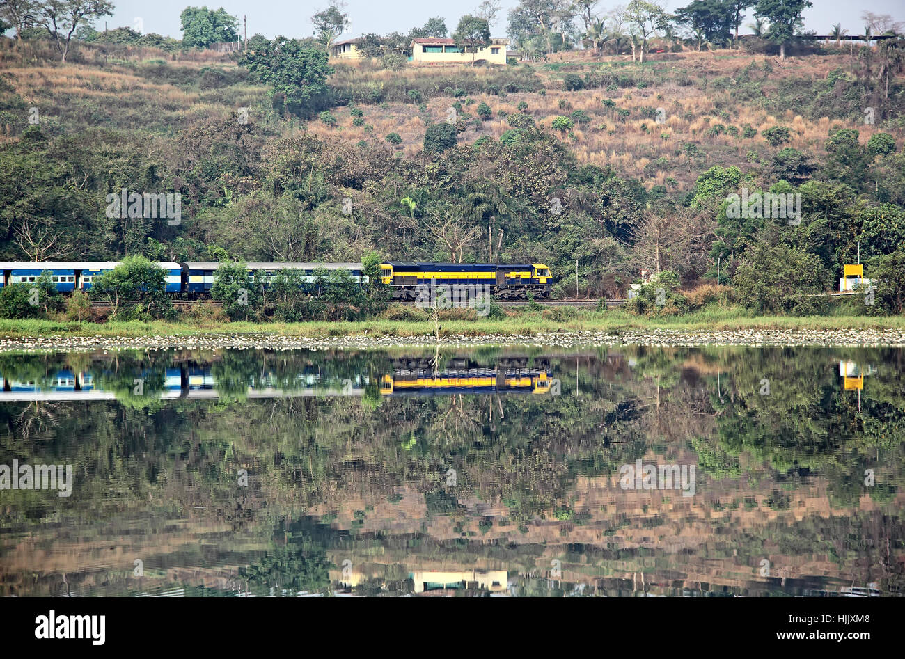 Treno passeggeri delle ferrovie indiane passando lungo le rive del lago in un terreno collinare, avvicinandosi ad una stazione in Goa, India Foto Stock