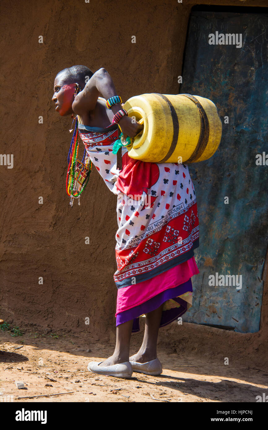 Donna Maasai alle prese con una canna di acqua sul retro, indossando costumi tradizionali, in un villaggio vicino al Masai Mara National Park, Kenya, Africa orientale Foto Stock