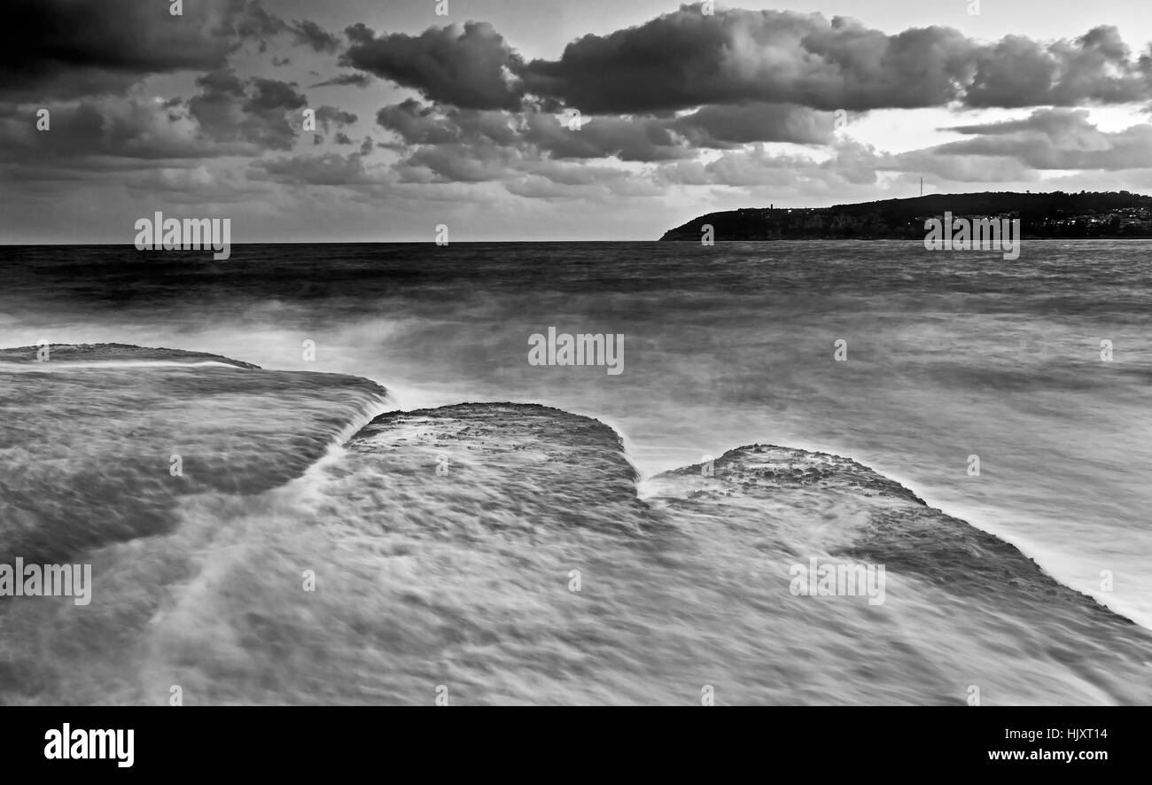 Rocce piatte sorvolati da un forte flusso di onda in arrivo alla spiaggia di acqua dolce delle spiagge del Nord di Sydney, Australia. Sunset seascape convertito in nero- Foto Stock