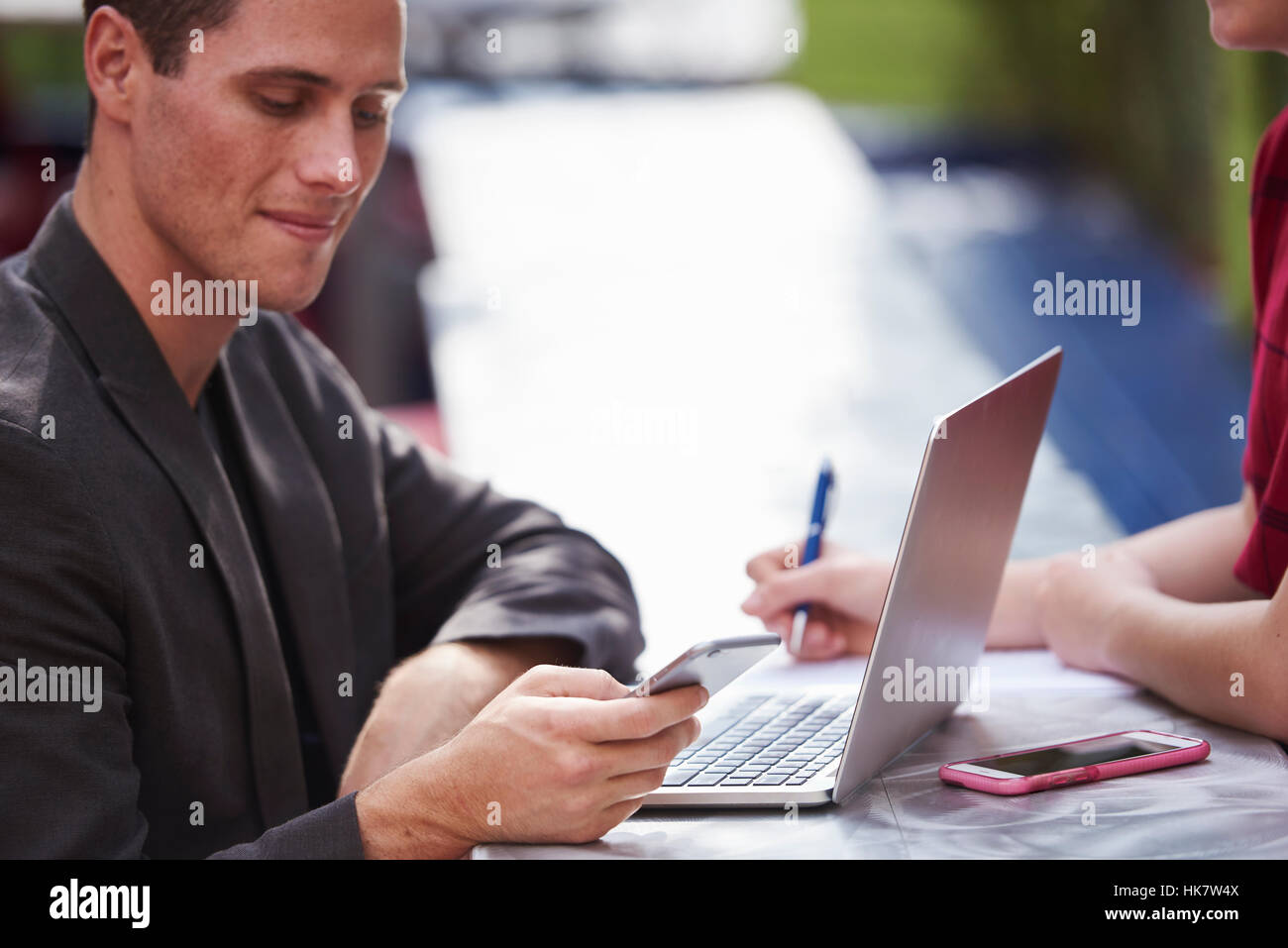 Un giovane uomo seduto ad un tavolo all'aperto con un computer portatile aperto che guarda un cellulare. Foto Stock