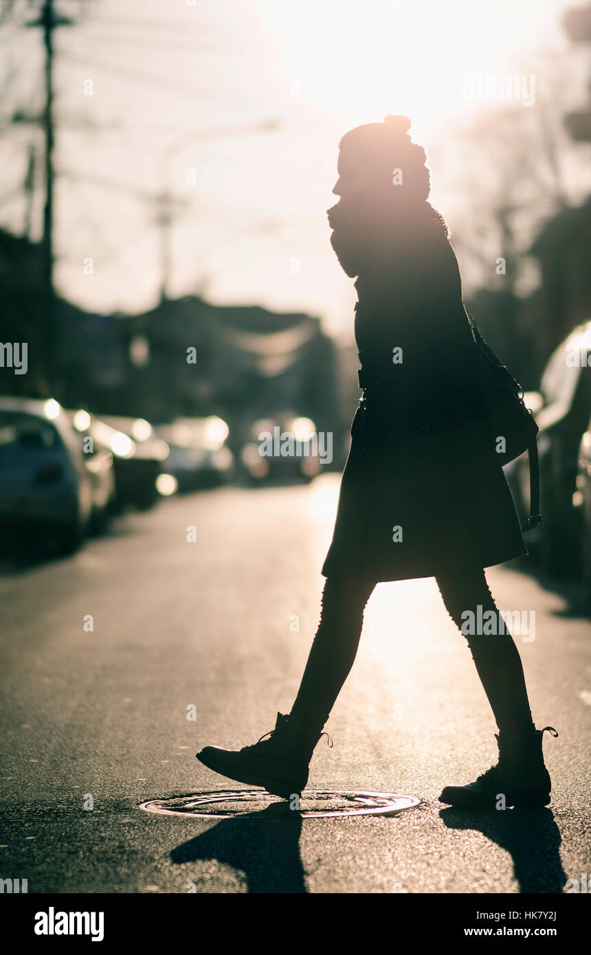 Una giovane donna in un mantello nero attraversando la strada in un freddo giorno al tramonto Foto Stock