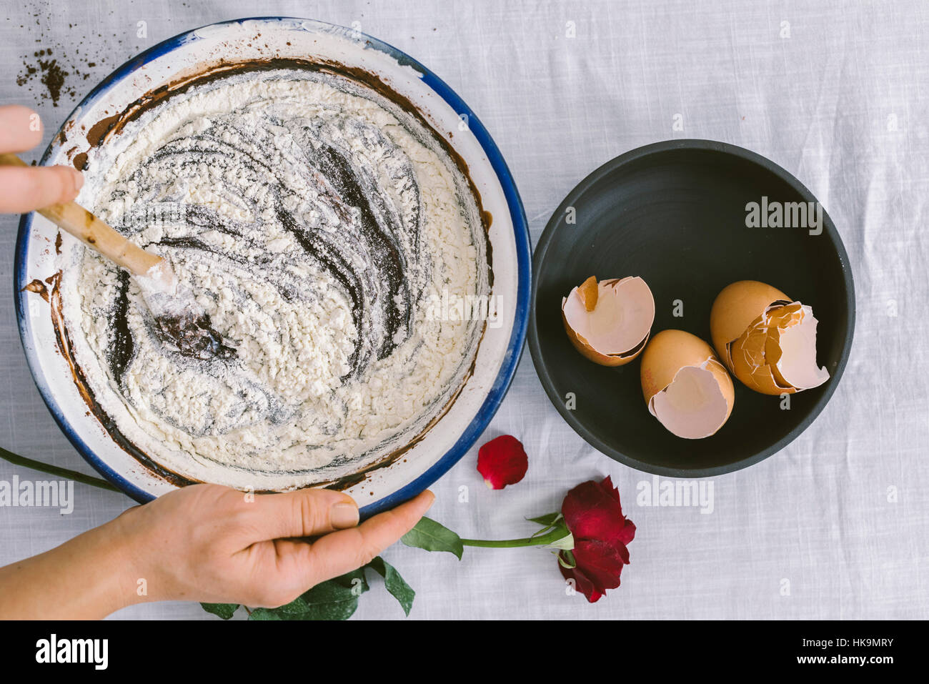 Le mani sotto agitazione una miscela di farina, cioccolato e cacao in polvere in un recipiente di miscelazione accompagnata dai gusci delle uova e una rosa rossa. Foto Stock