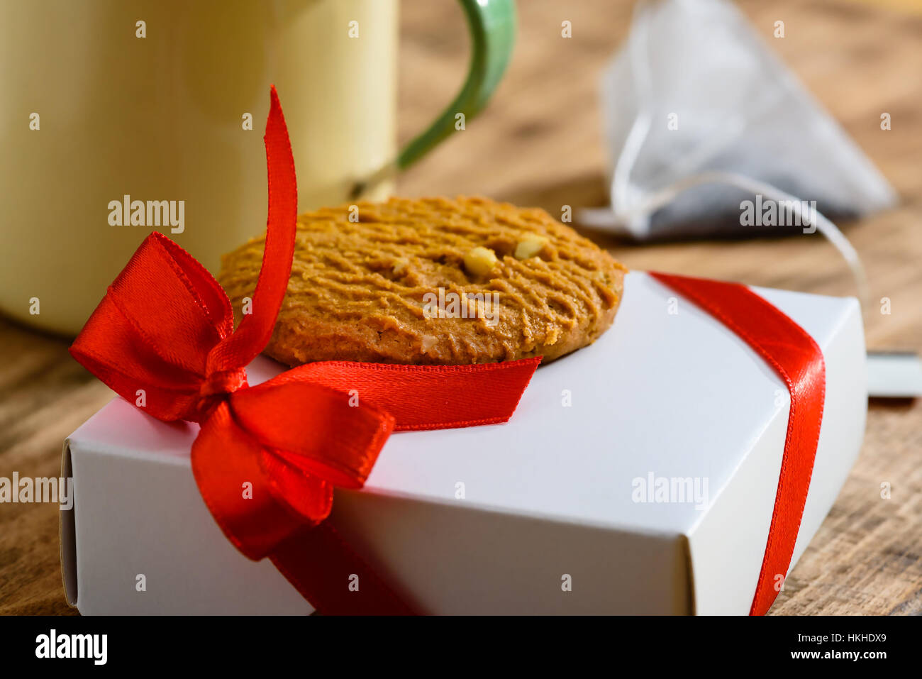 Piccolo presente di colore bianco con fiocco rosso e cookie sulla parte superiore insieme con parziale tazzina e bustine di the sul vassoio in legno Focus su cookie Foto Stock