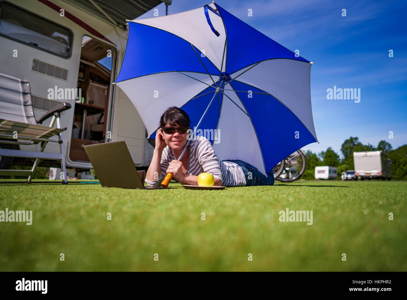 Donna sull'erba, guardando il laptop sotto ombrellone vicino al сamping. Caravan auto vacanza. Vacanza per la famiglia in viaggio, viaggio vacanza in camper Foto Stock