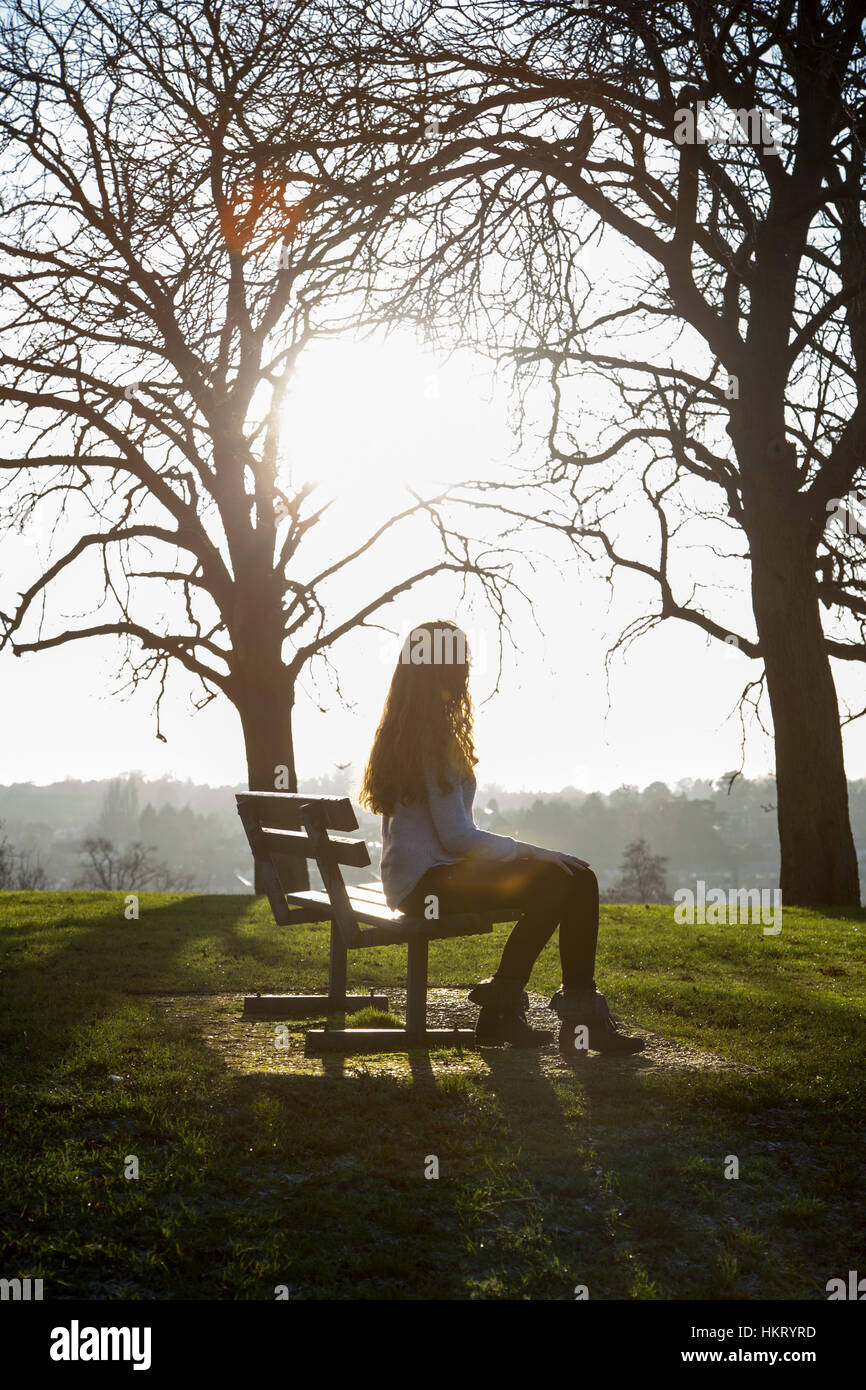 Donna in venti con capelli lunghi, seduta su una panchina nel parco guardando l'orizzonte, all'aperto, silhouette. Foto Stock