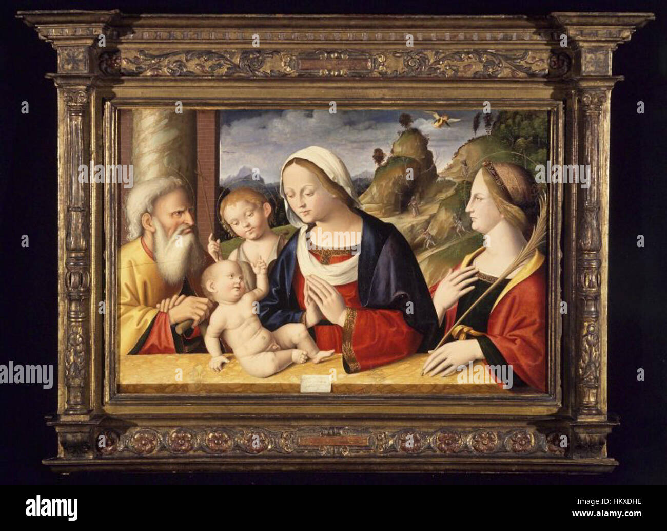 La *Sacra famiglia con i Santi Giovanni Battista e Caterina d'Alessandria* di Marco Palmezzano è un dipinto religioso che raffigura la Sacra famiglia con due santi. L'opera d'arte è un esempio di arte religiosa rinascimentale, che mescola colori vivaci con soggetti divini e offre una narrazione visiva della fede cristiana. Foto Stock
