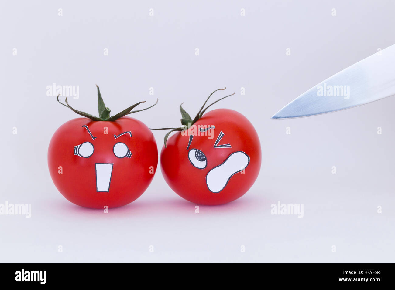 Due freschi pomodori rossi con paura volti e una lama affilata lama nella parte anteriore del fondo bianco Foto Stock