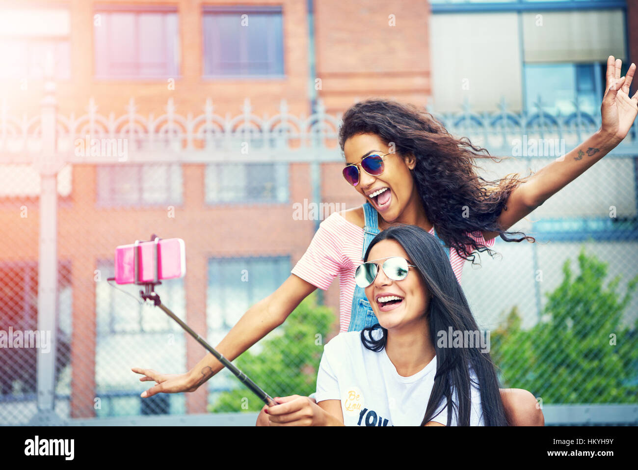 Due esuberanti amiche compiendo gesti esagerata tenendo un autoritratto al di fuori con il rosa telefono cellulare collegato al polo stick Foto Stock