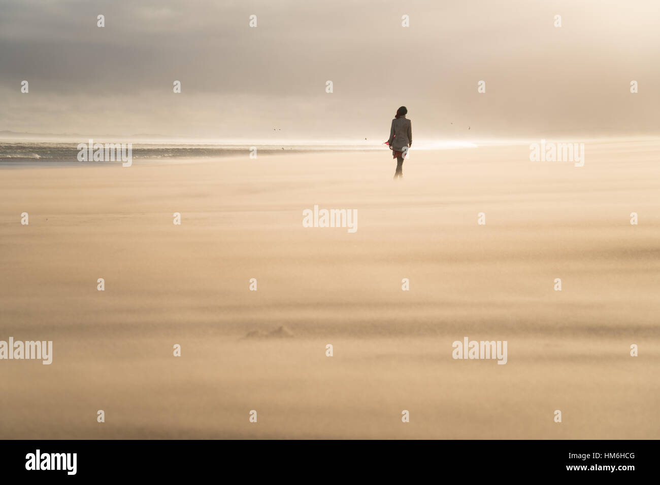 Unica donna a camminare su lonely spiaggia sabbiosa retroilluminata con beret hat e il mare lontano e onde Foto Stock
