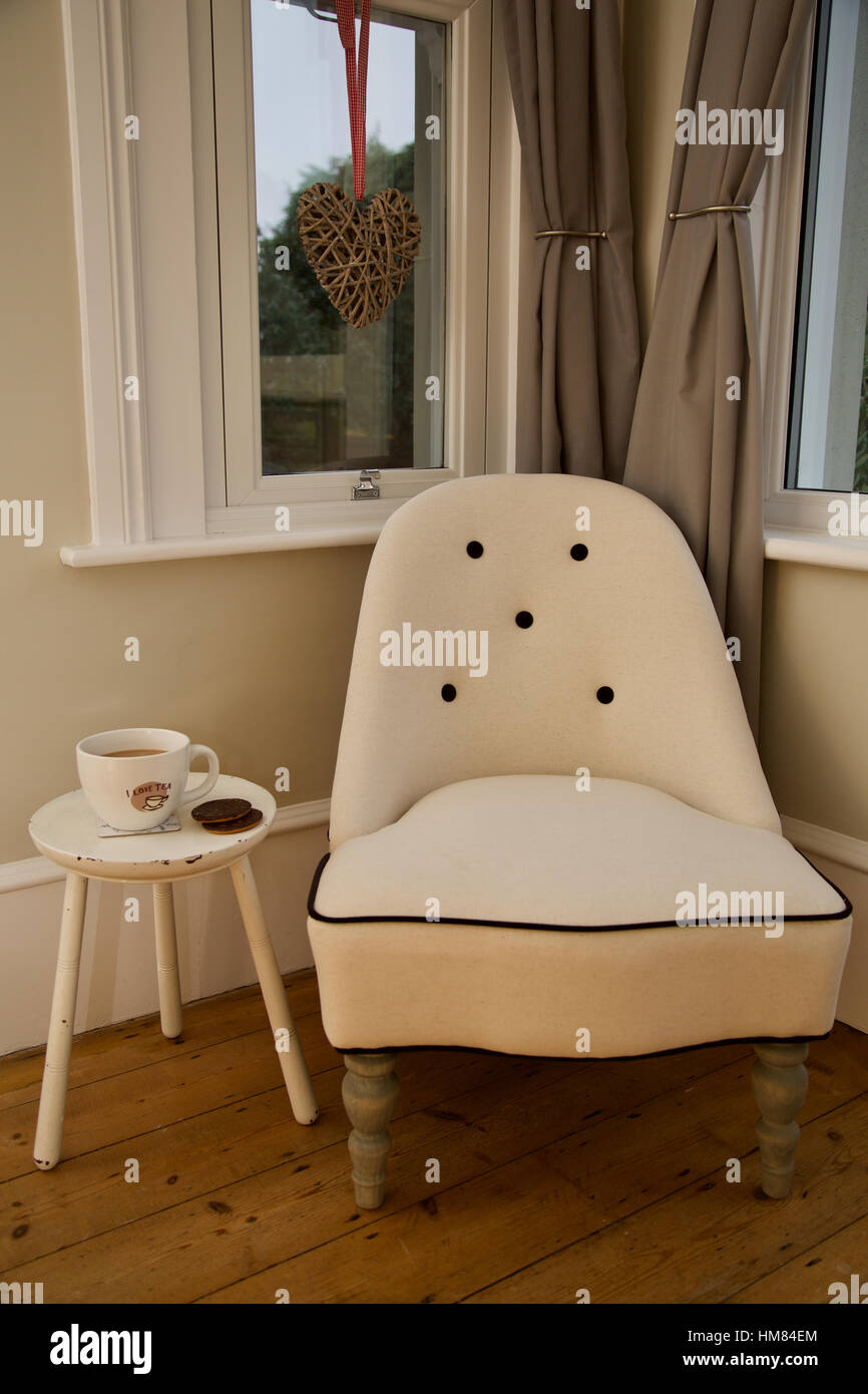 Vittoriano tradizionale sedia crema nell'angolo dalla finestra a casa con la tabella con la tazza di tè e biscotti pronti a prendersi una pausa di relax Foto Stock