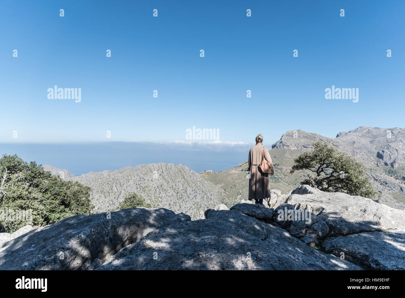 Persona di sesso femminile con un lungo soprabito e borsetta in piedi sulla cima della montagna sotto il cielo blu Foto Stock