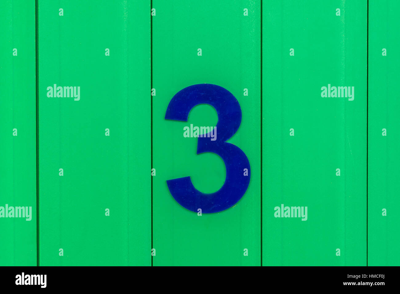 Il numero tre (3), in blu, su una superficie di colore verde che è fatta di legno Foto Stock