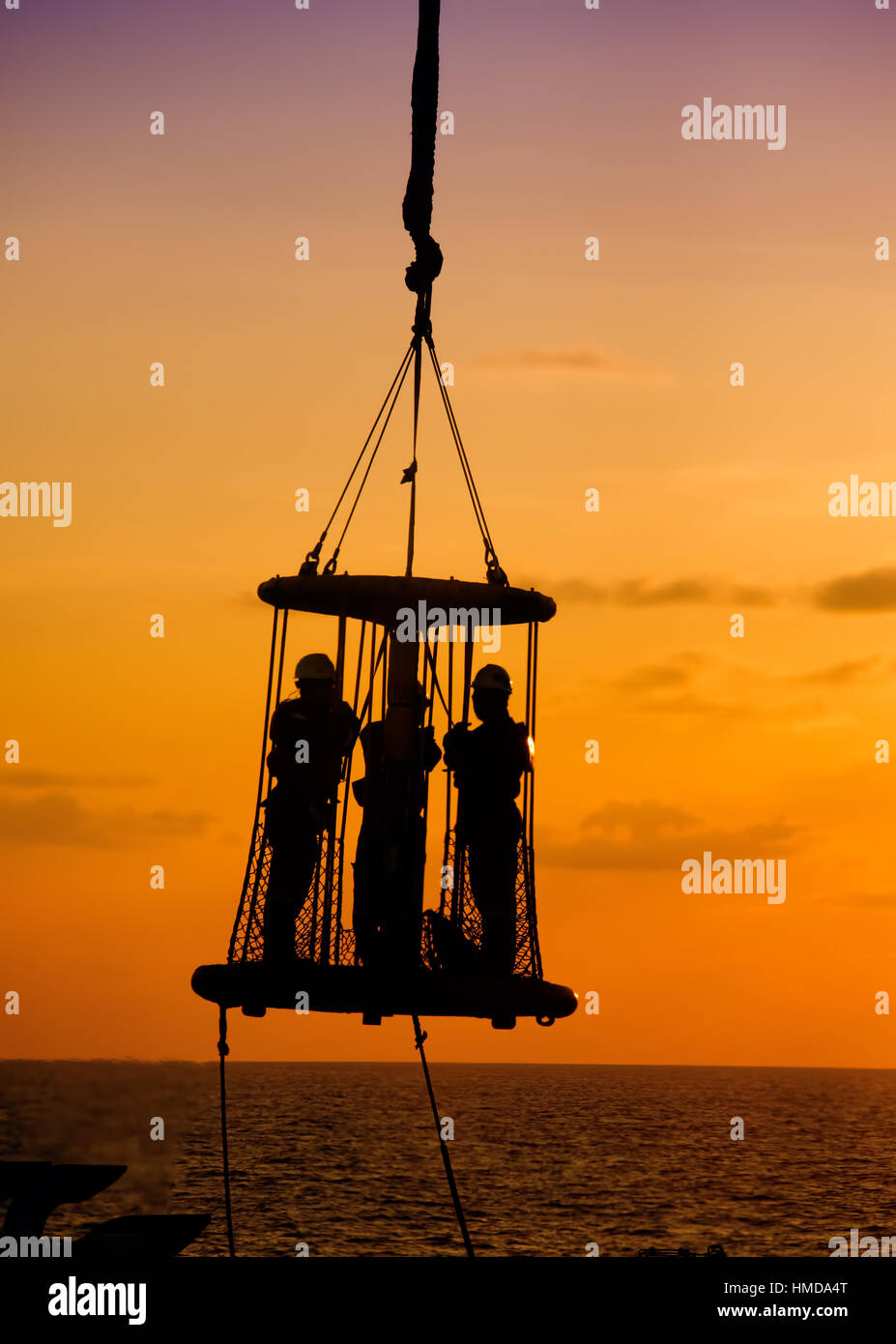 Silhouette lavoratore offshore appeso all'interno di sicurezza personale cestello in mare durante il tramonto Foto Stock