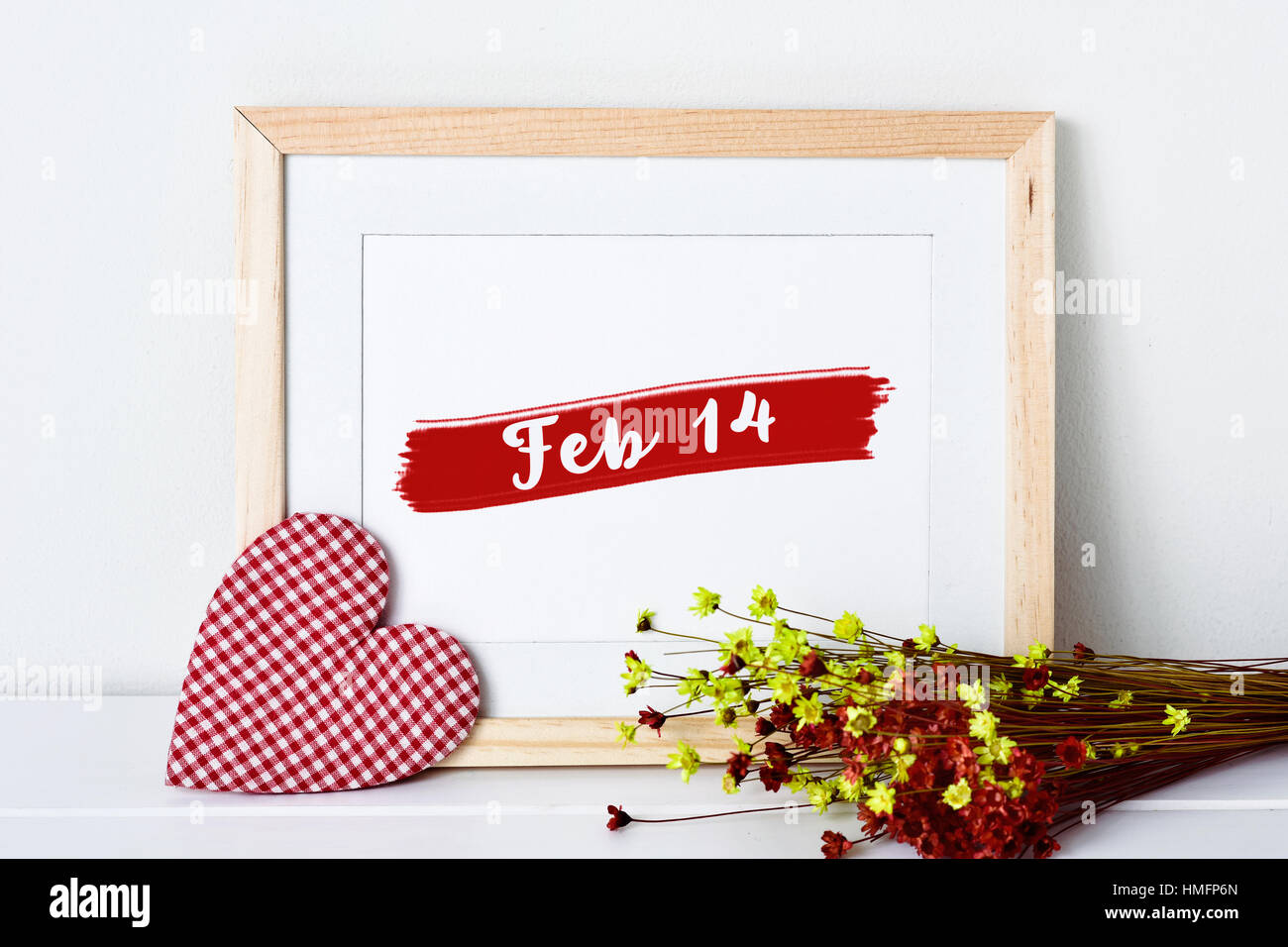 Un tessuto del cuore, una tavola di legno con cornice foto con il testo feb 14 e un mazzetto di rosso e di giallo fiori secchi su una superficie bianca Foto Stock