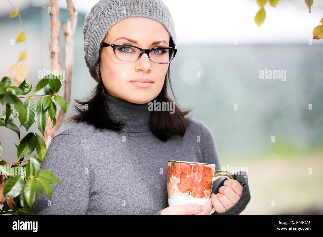 La splendida green eyed giovane donna in abiti caldi, visione di occhiali e Grey Hat sorridente, bere il tè in una grande tazza all'aperto nel periodo autunnale Foto Stock