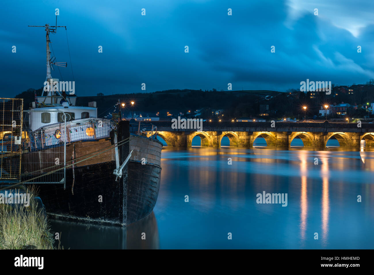 Una casa galleggiante si trova sulla riva del fiume Torridge in Bideford come le luci sul lungo ponte si accende al crepuscolo. Foto Stock