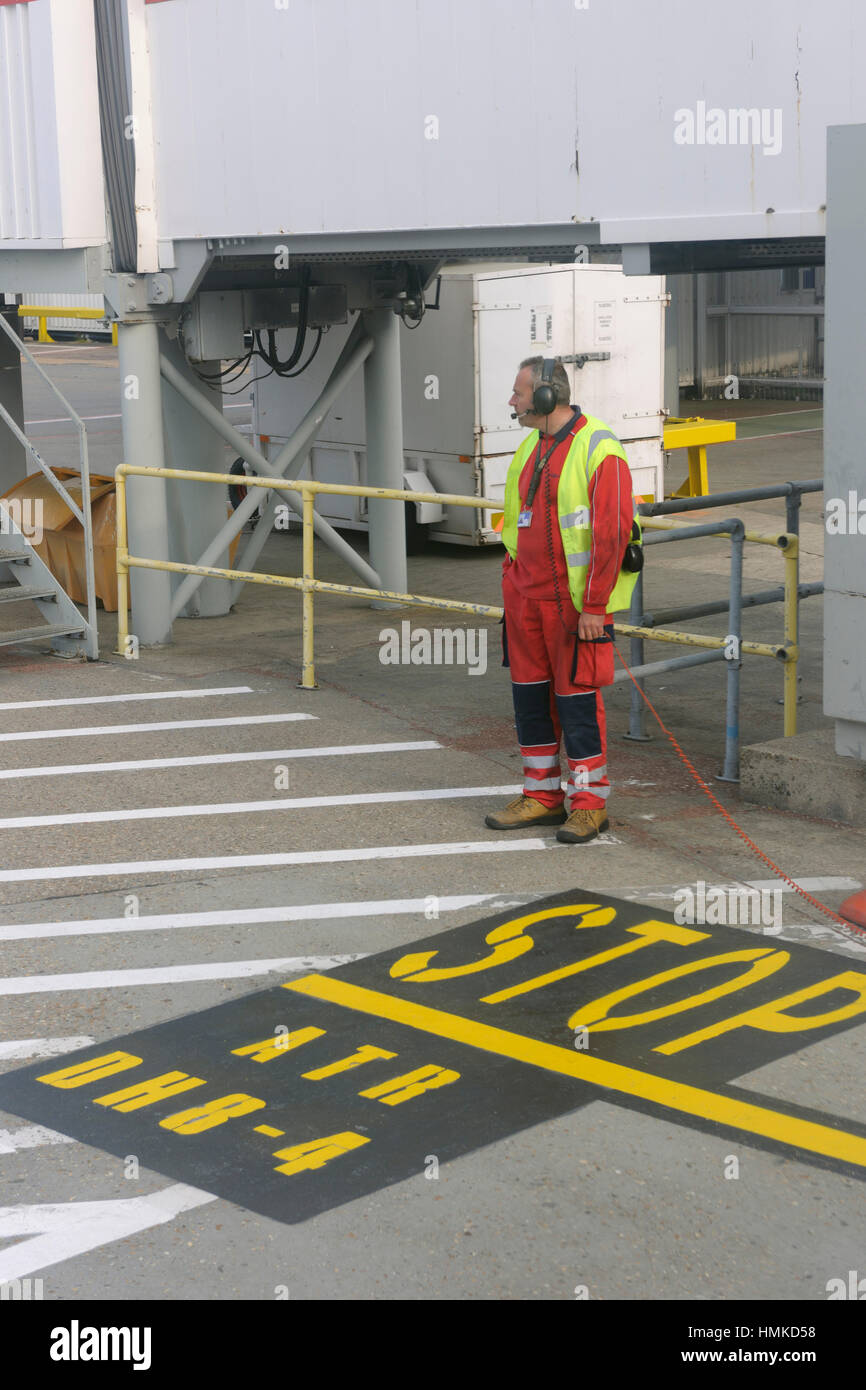 Uomo che indossa la cuffia con arancio e giallo ad alta viz tabard in piedi sul parcheggio stop contrassegni per DHC8 Q400 e ATR Foto Stock