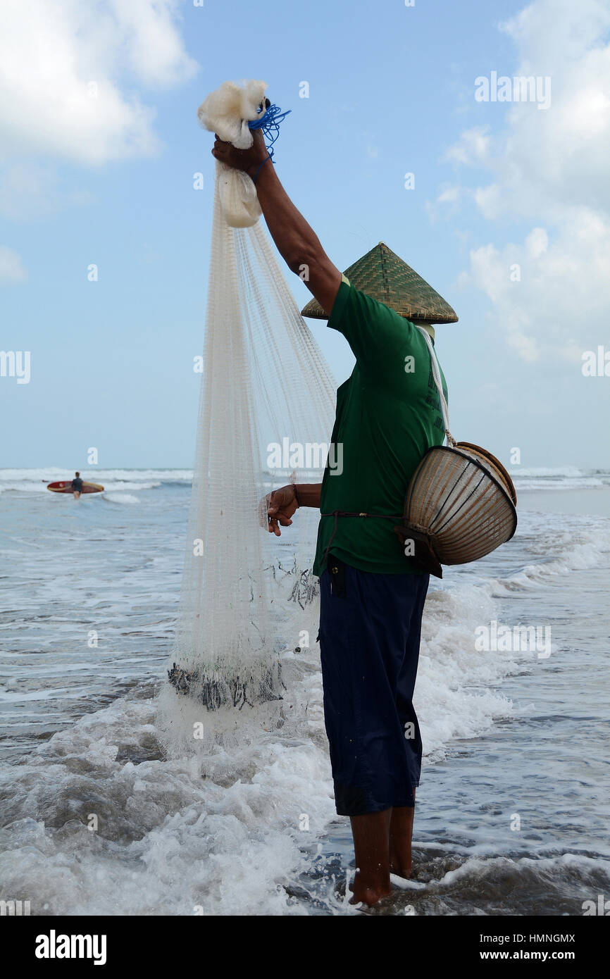 Un pescatore sulla spiaggia di Seminyak raccoglie la sua pesca mentre un surfista osserva lui Foto Stock