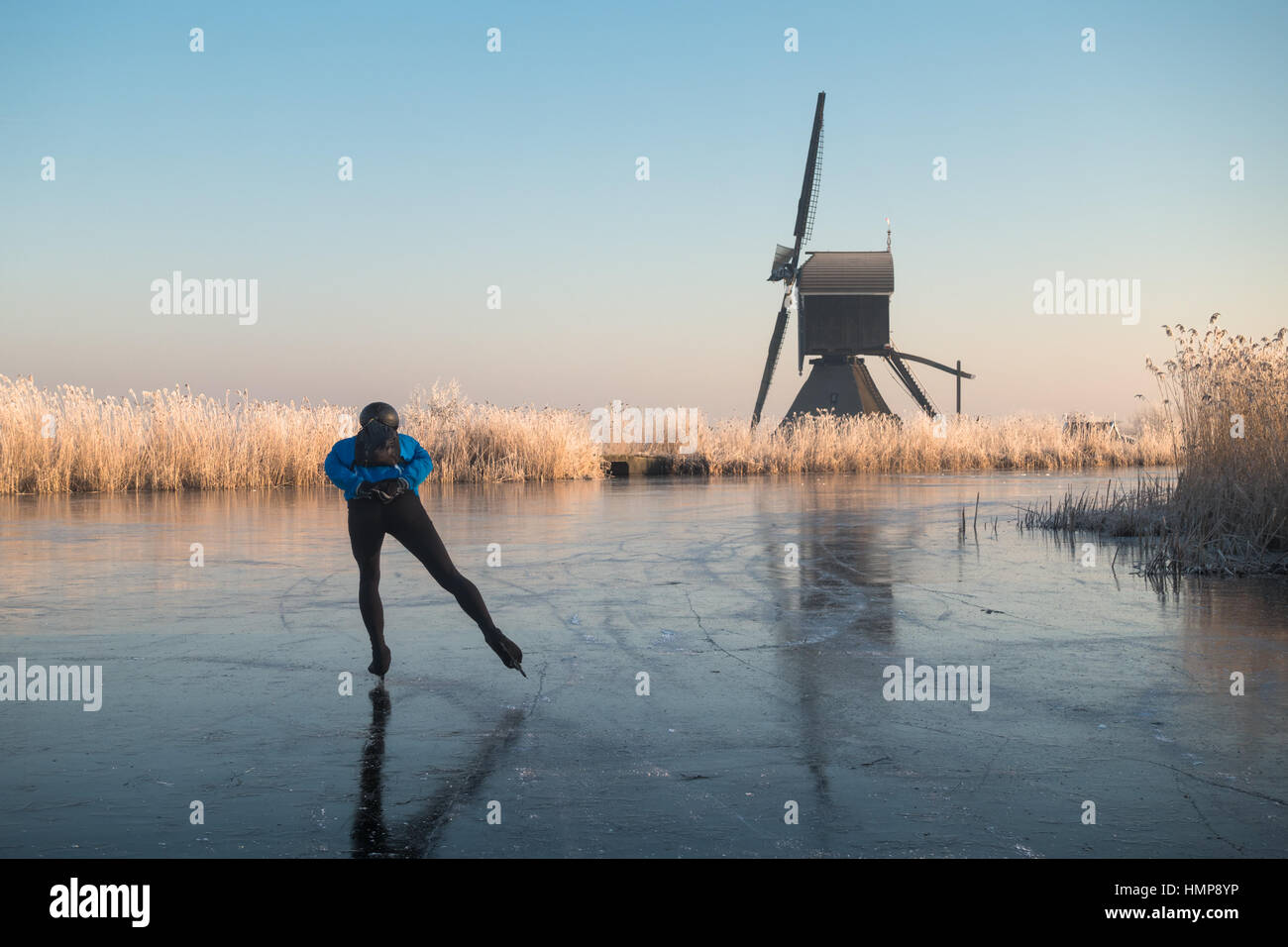 Persona pattinaggio sul ghiaccio da soli su un fiume congelato passato un mulino a vento e trasformata per forte gradiente smerigliati canneti a Kinderdijk in inverno nei Paesi Bassi. Foto Stock