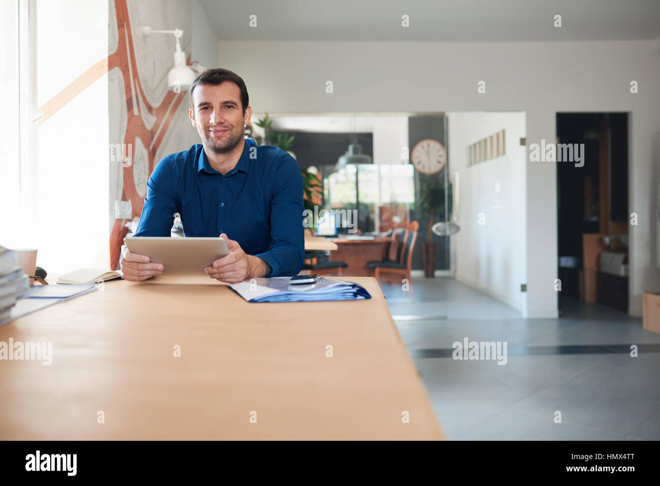 Ritratto di un fiducioso e imprenditore di successo che indossa un collare aperto shirt seduto alla sua scrivania utilizzando una tavoletta digitale nel suo ufficio moderno Foto Stock