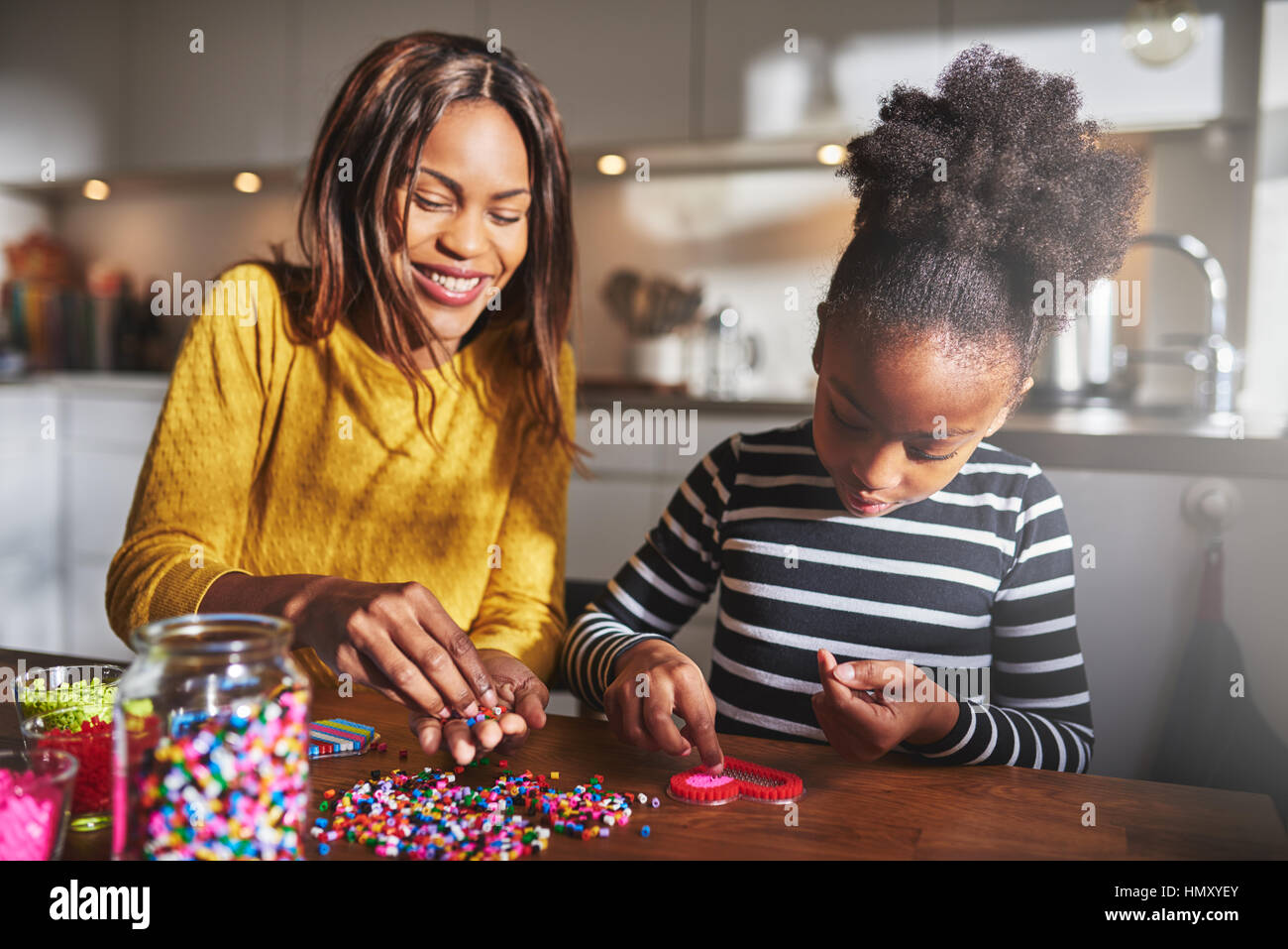 Attraente giovane americano africano genitore femminile aiutando carino bambino in maglia a strisce di scegliere i colori del tallone per la forma di cuore il telaio sul tavolo in cucina Foto Stock