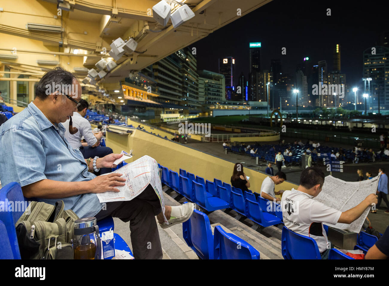 Giocatore cinese di tabacco da fumo tubo e leggere il giornale e altre persone all'Ippodromo Happy Valley di Hong Kong, Cina, la sera. Foto Stock