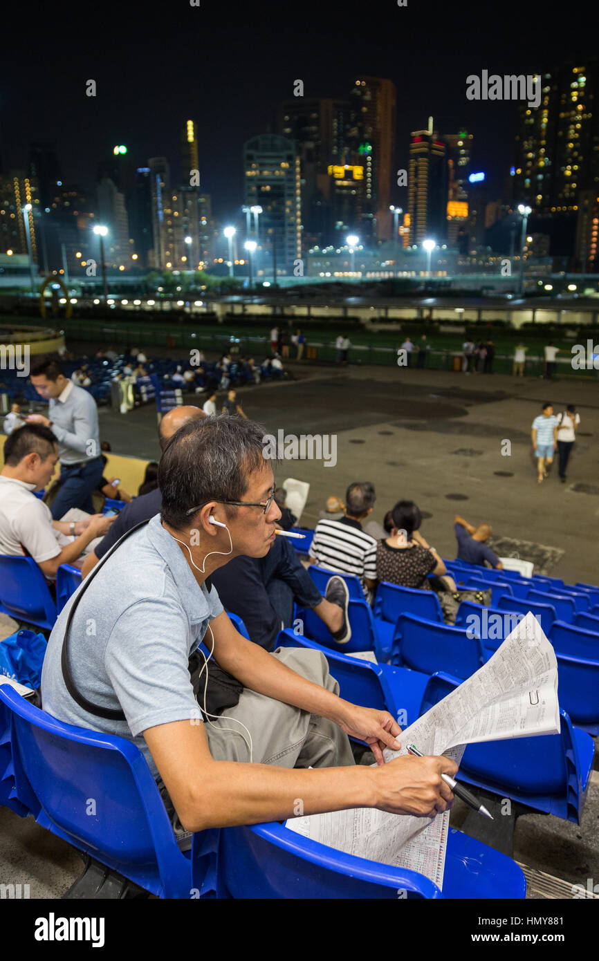 Giocatore cinese il fumo di sigarette e di leggere il giornale e altre persone all'Ippodromo Happy Valley di Hong Kong, Cina, la sera. Foto Stock