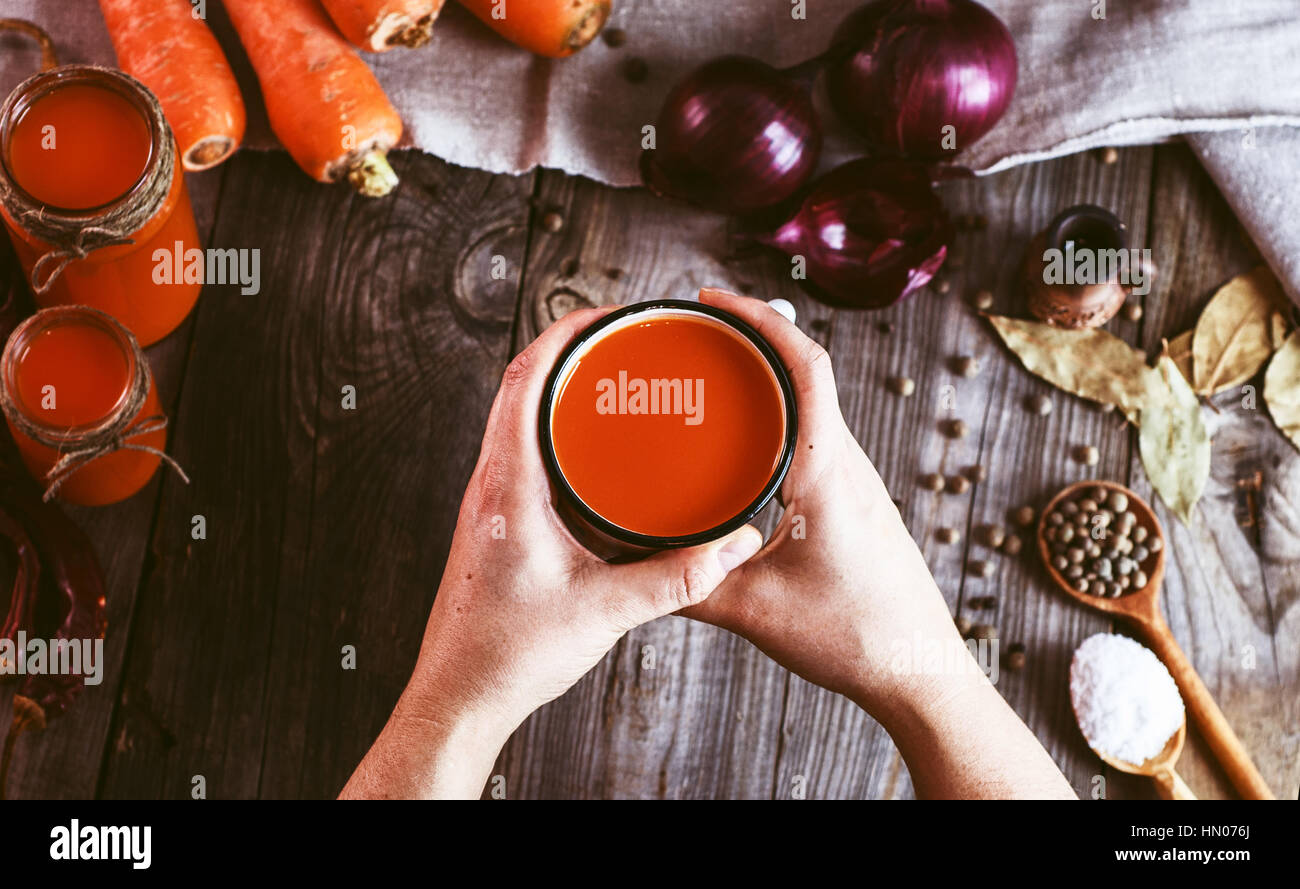 Due mani femminili di trattenimento di un ferro da stiro la tazza con il succo di carota sullo sfondo della tabella con verdure fresche e spezie Foto Stock