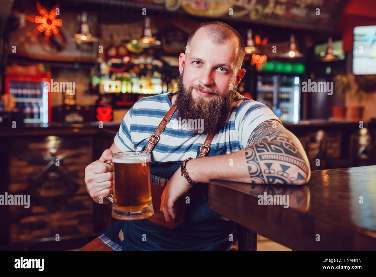Barman Barbuto con tatuaggi di indossare un grembiule seduti al bar e in mano un bicchiere di birra Foto Stock