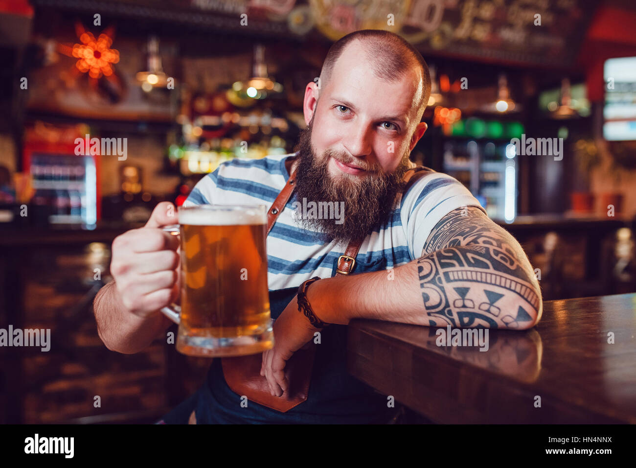 Barman Barbuto con tatuaggi di indossare un grembiule seduti al bar e in mano un bicchiere di birra Foto Stock