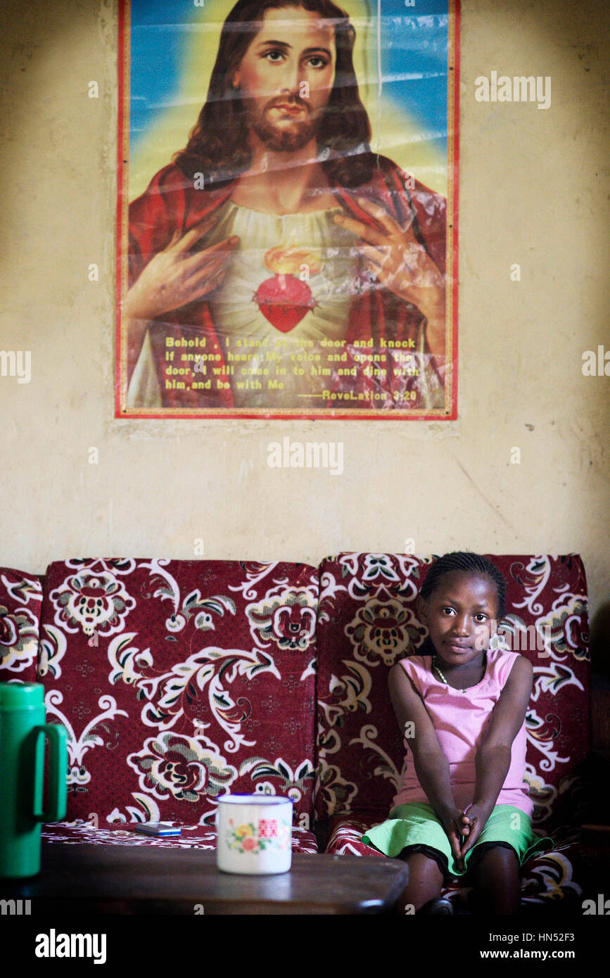 Giovane ragazza ugandese si siede su una sedia sotto un poster di Gesù Cristo nella sua casa di famiglia Foto Stock