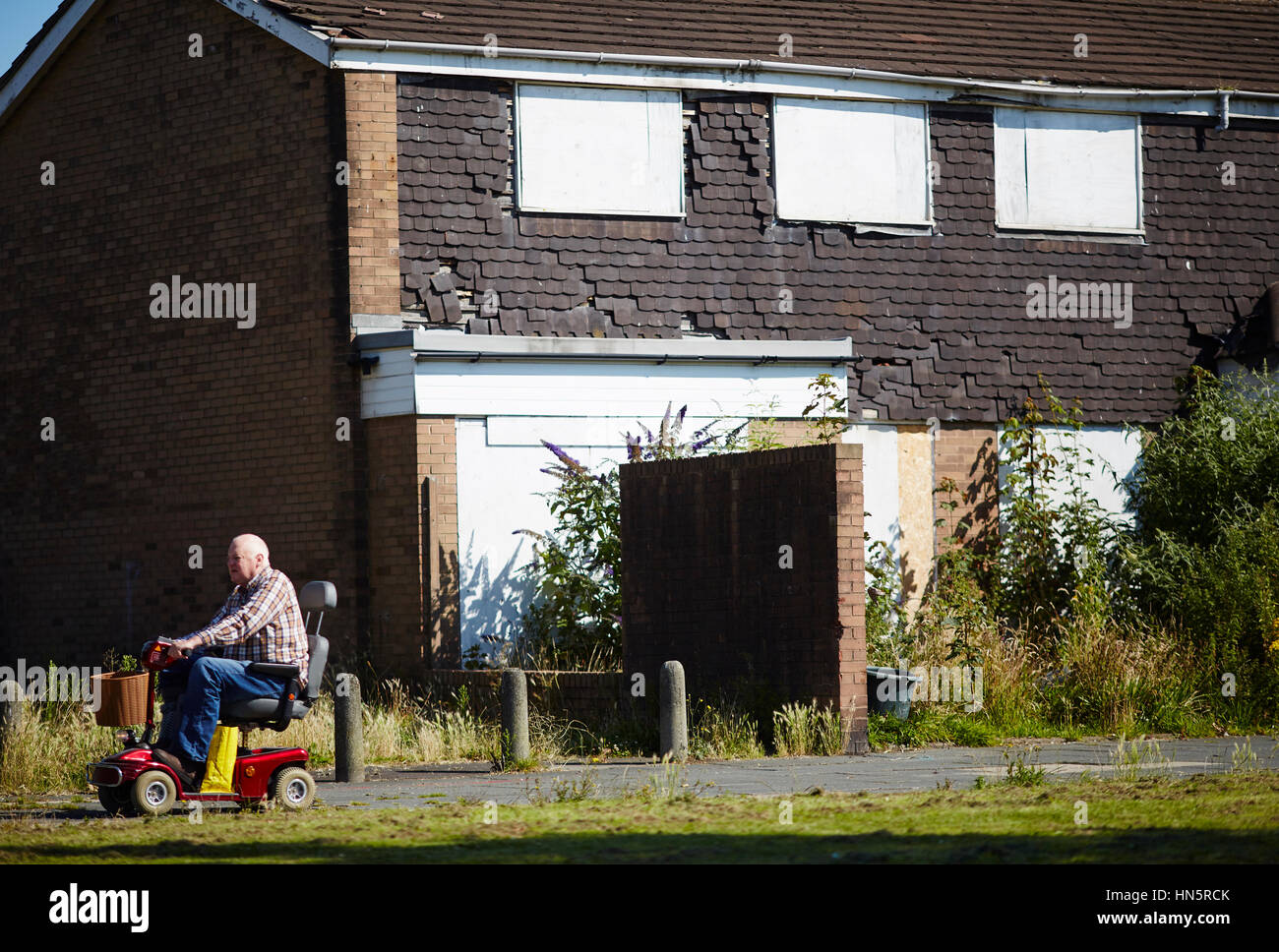 Vecchio sovrappeso uomo grasso sulla mobilità scooter passando vecchio alloggiamento magazzino malandato imbarcati su Windows e il vuoto in East Manchester gioco di eseguire dow Foto Stock