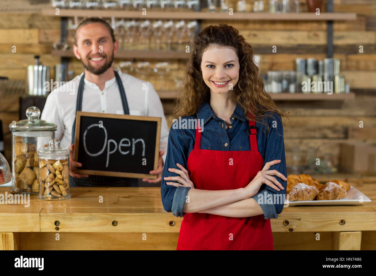 Ritratto di cameriera in piedi con le braccia incrociate al contatore in cafÃƒÂ© con cameriere tenendo aperte segno bordo in background Foto Stock