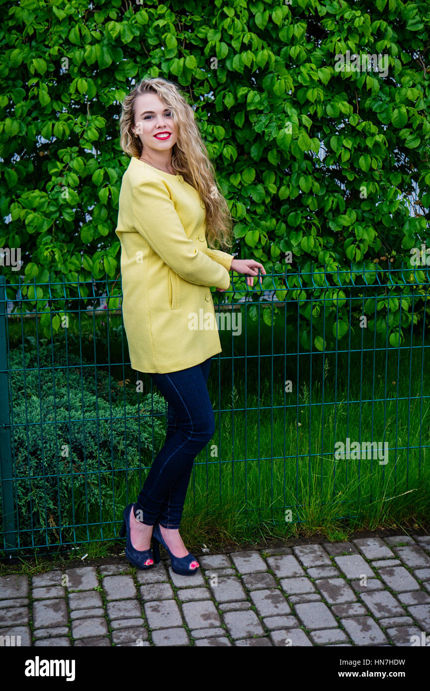 Ritratto di attraente ragazza bionda con capelli ondulati, emotivamente sorridente e ponendo in telecamera. Indossando rivestimento giallo. In piedi contro albero verde. A piedi Foto Stock