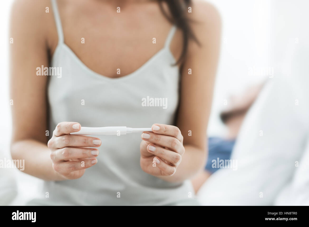 Giovane donna seduta sul letto e in attesa di un test di gravidanza di risultati, il suo fidanzato dorme sullo sfondo Foto Stock