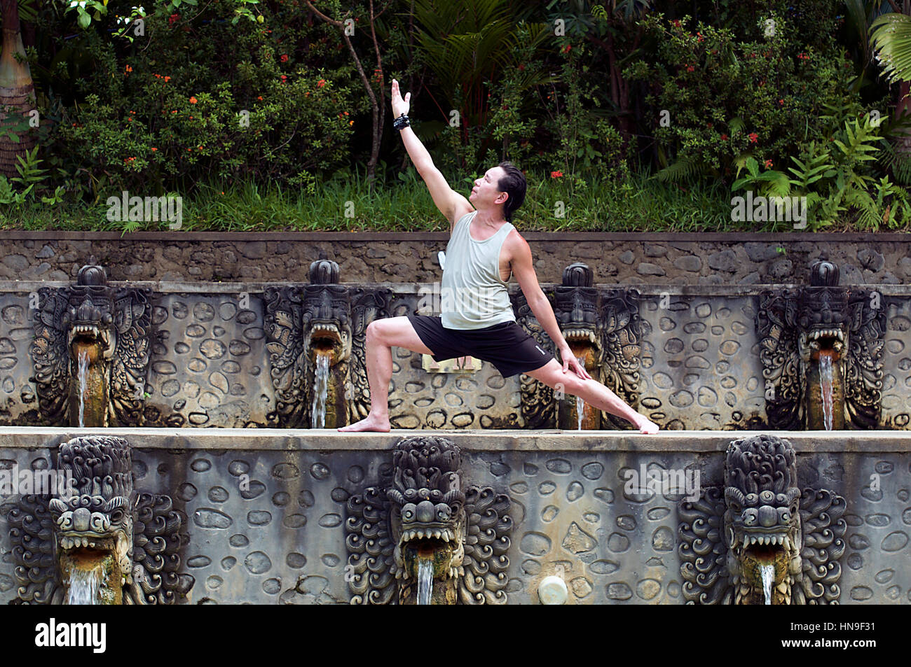 Middle-Aged Uomo asiatico detiene un guerriero di Hatha Yoga pone a Banjar Hot Springs per piscine di Bali, Indonesia come acqua si riversa da creature Dragon-Like' bocche Foto Stock