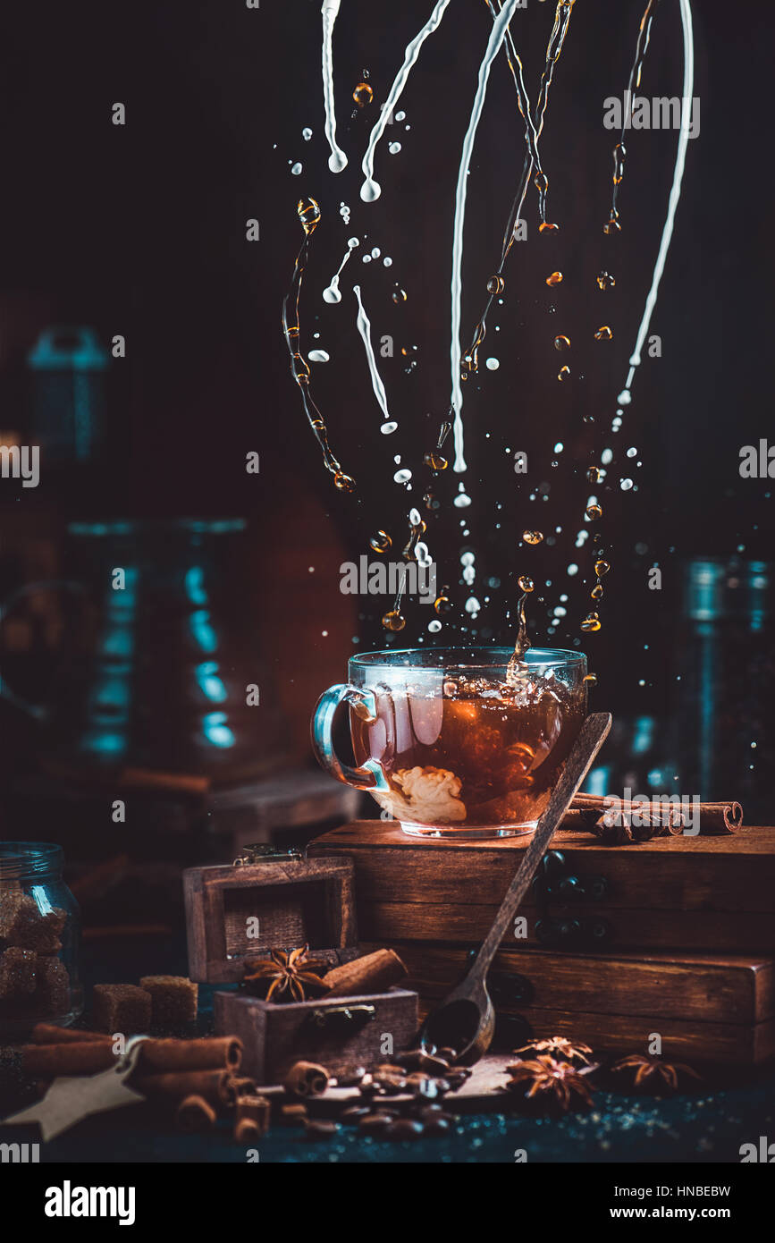 Latte di ritorcitura e schizzi di caffè in una coppa trasparente su sfondo scuro con macinino da caffè, cucchiai, cannella e chicchi di caffè Foto Stock