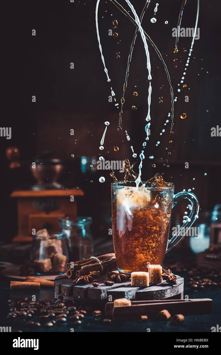Latte di ritorcitura e schizzi di caffè in una coppa trasparente su sfondo scuro con macinino da caffè, cucchiai, cannella e chicchi di caffè Foto Stock