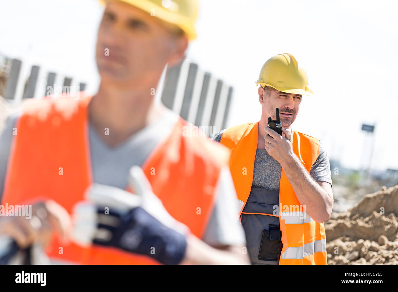 Di mezza età lavoratore maschio utilizzando un walkie-talkie con il collega in primo piano a sito in costruzione Foto Stock