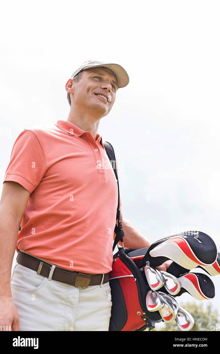 Felice uomo di mezza età che guarda lontano mentre trasportano borsa da golf contro il cielo chiaro Foto Stock