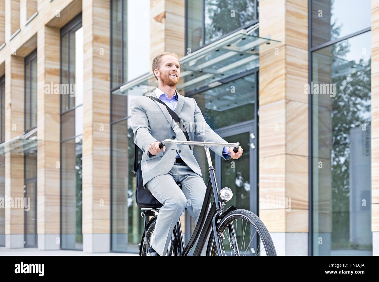 Imprenditore sorridente bicicletta equitazione all'esterno dell'edificio Foto Stock