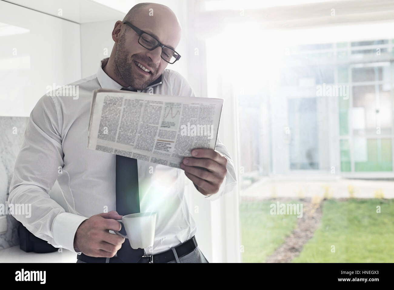 Adulto medio imprenditore su chiamata mentre si tiene il giornale e tazza di caffè a casa Foto Stock