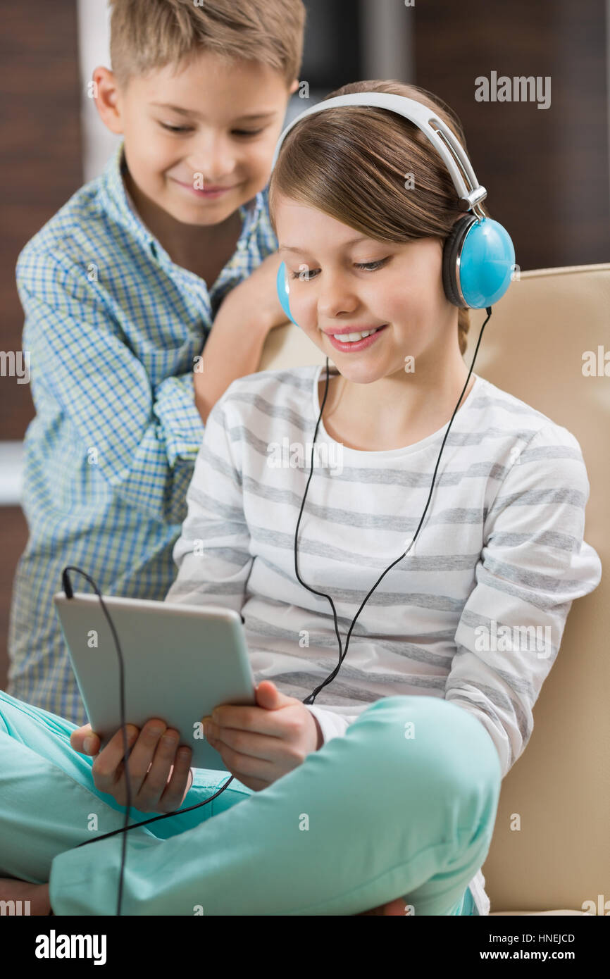 Ragazza carina ascoltando musica su tavoletta digitale mentre il fratello in piedi dietro di lei a casa Foto Stock