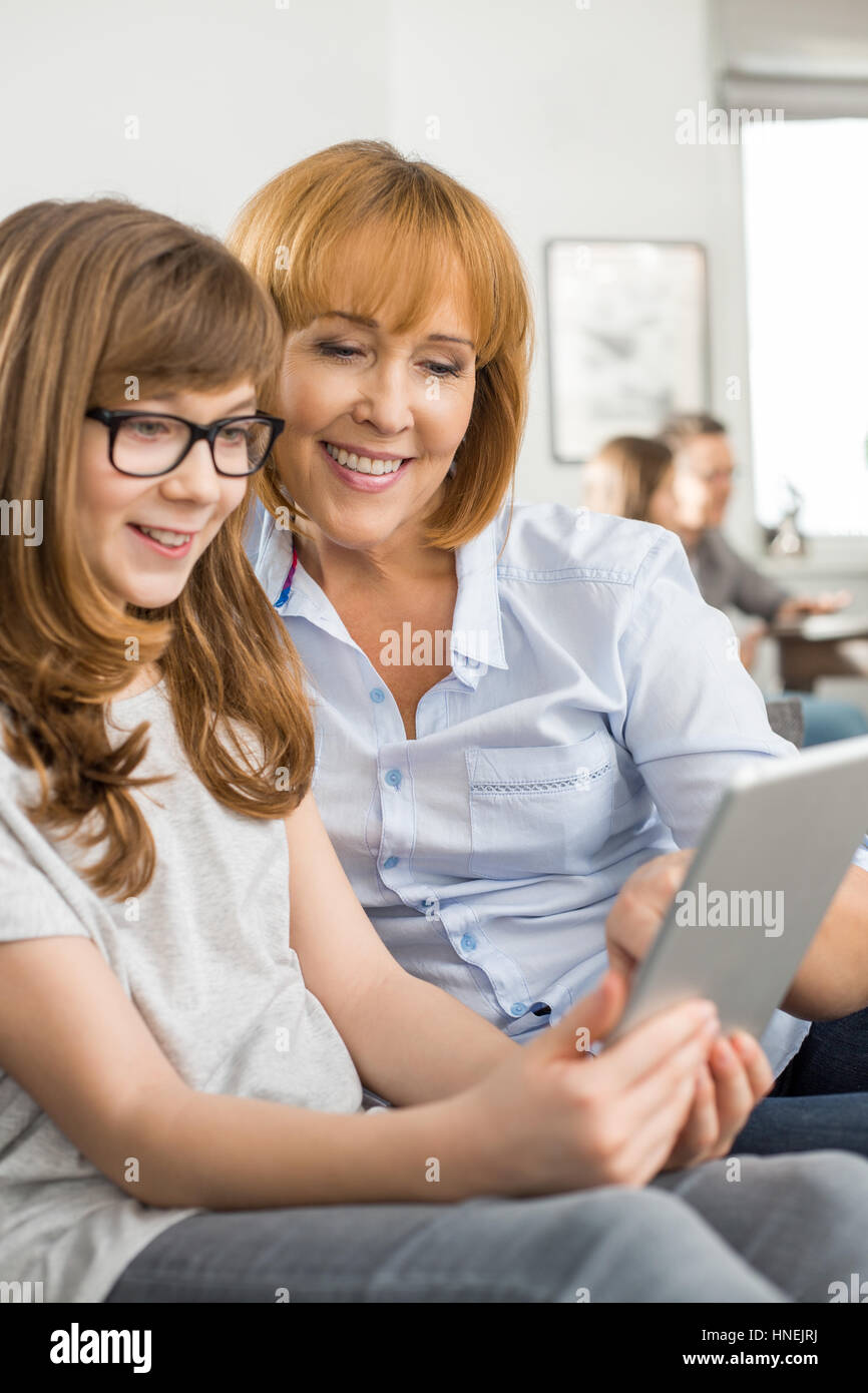 Felice madre e figlia utilizzando tablet PC con la famiglia seduta in background a casa Foto Stock