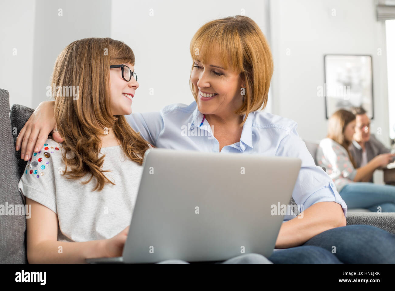 Felice madre e figlia con il computer portatile mentre la famiglia seduta in background a casa Foto Stock