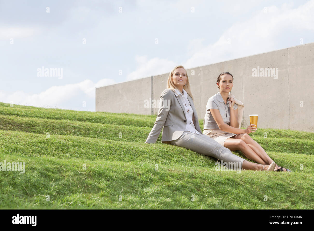A piena lunghezza rilassata imprenditrici in formali seduti sulle fasi di erba contro sky Foto Stock